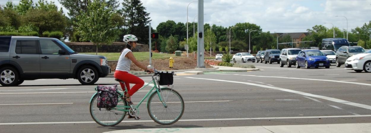 Woman on a bicycle waits at a stoplight. There is a truck in the travel lane to her left.