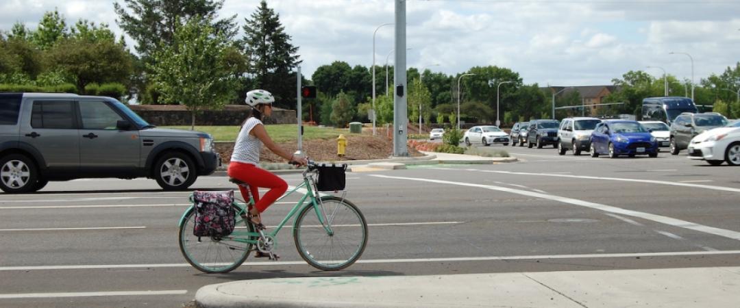 Woman on a bicycle waits at a stoplight. There is a truck in the travel lane to her left.